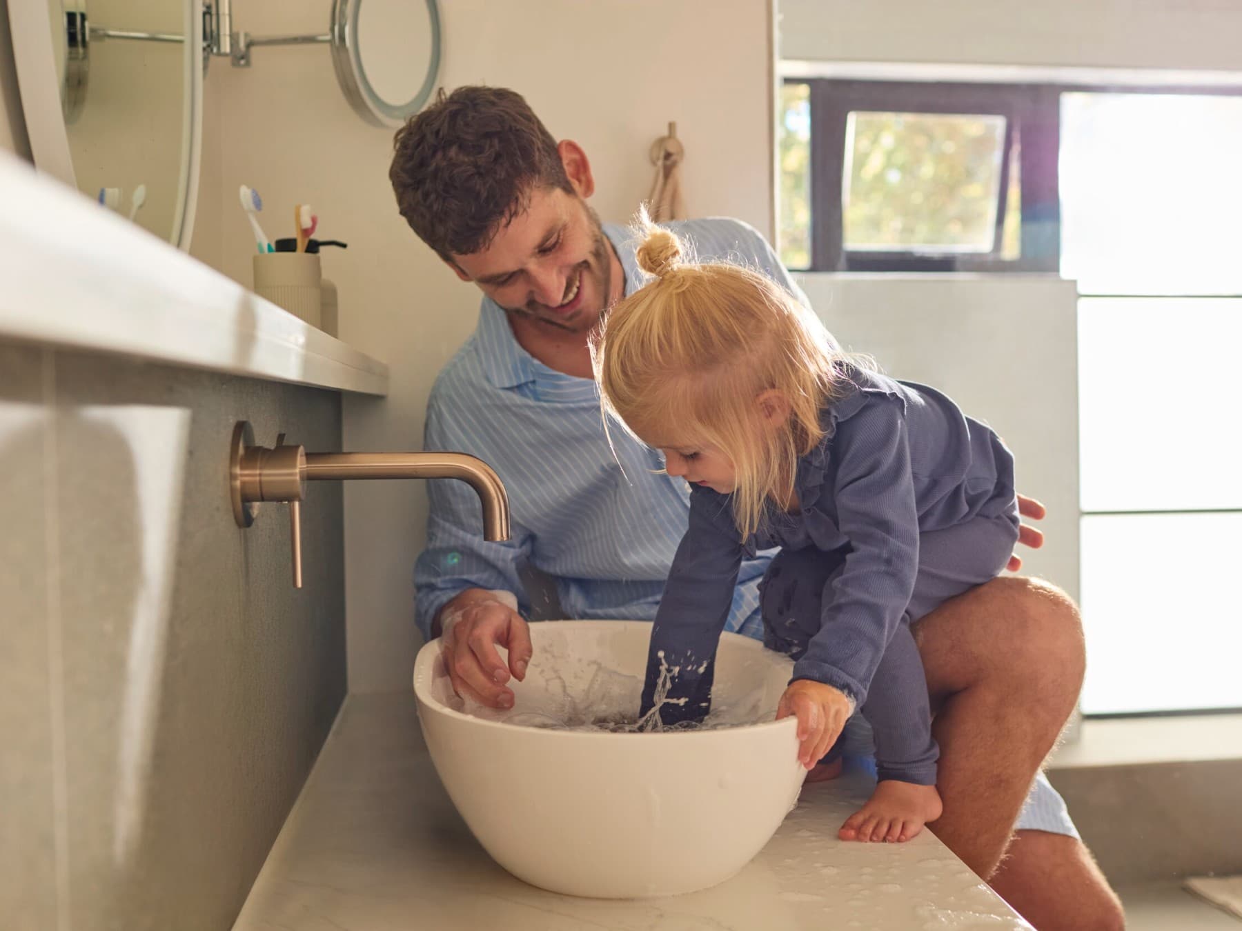 Татко и ќерка стојат пред мијалник во бањата и си играат со водата.