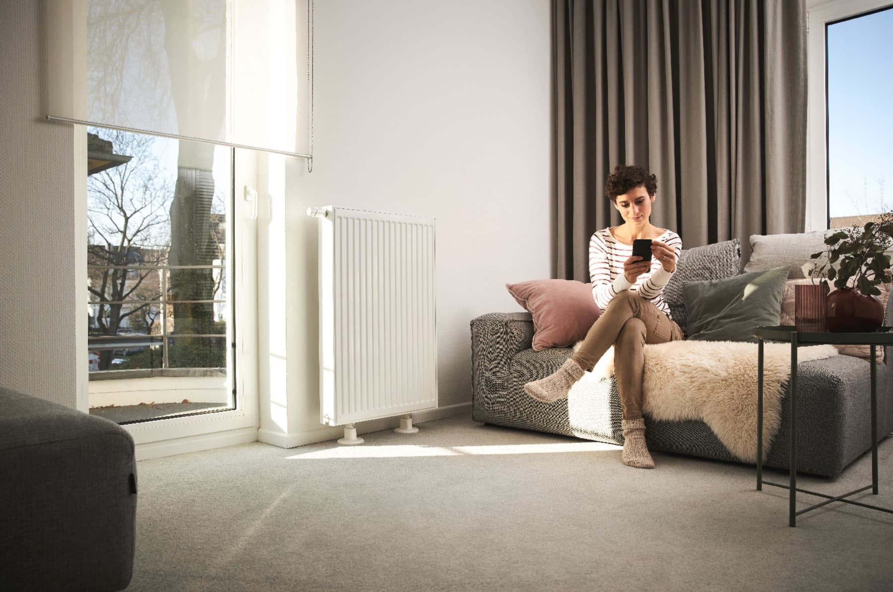 Жена седи на софа до радијатор гледајќи во мобилен телефон