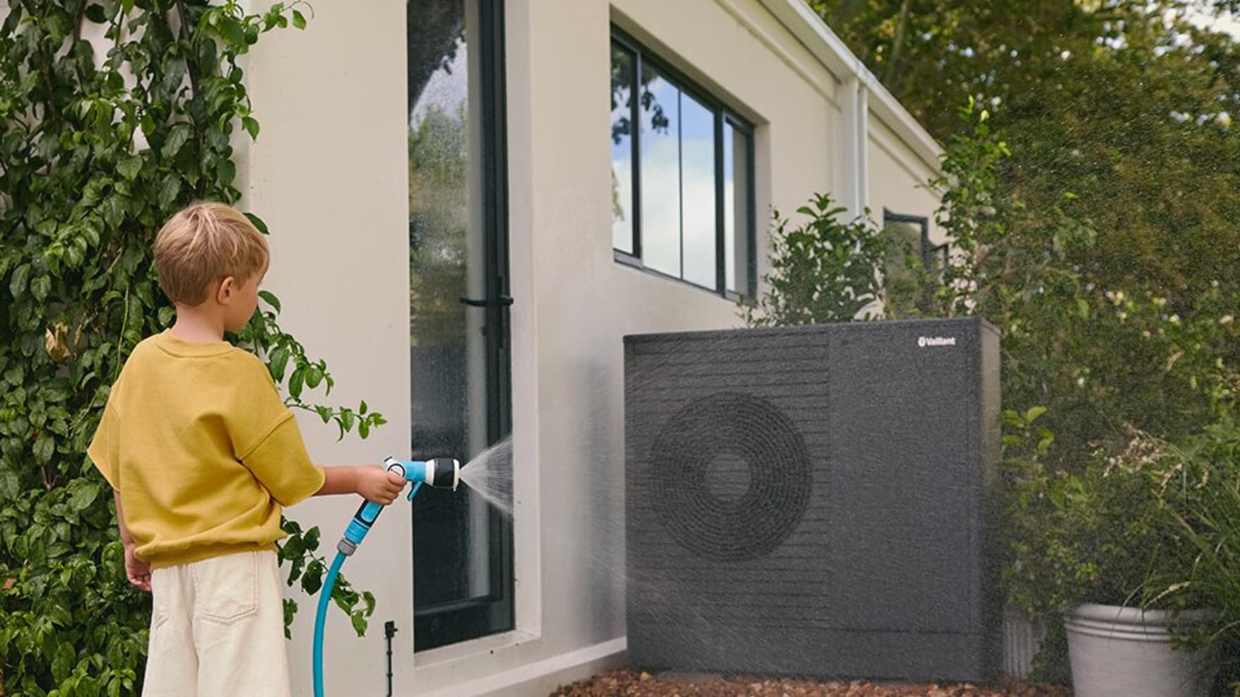 Момче прска вода врз топлинската пумпа со градинарското црево