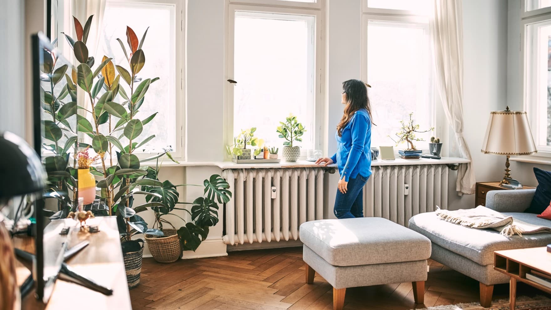 Жена стои во дневната соба пред прозорците и гледа надвор