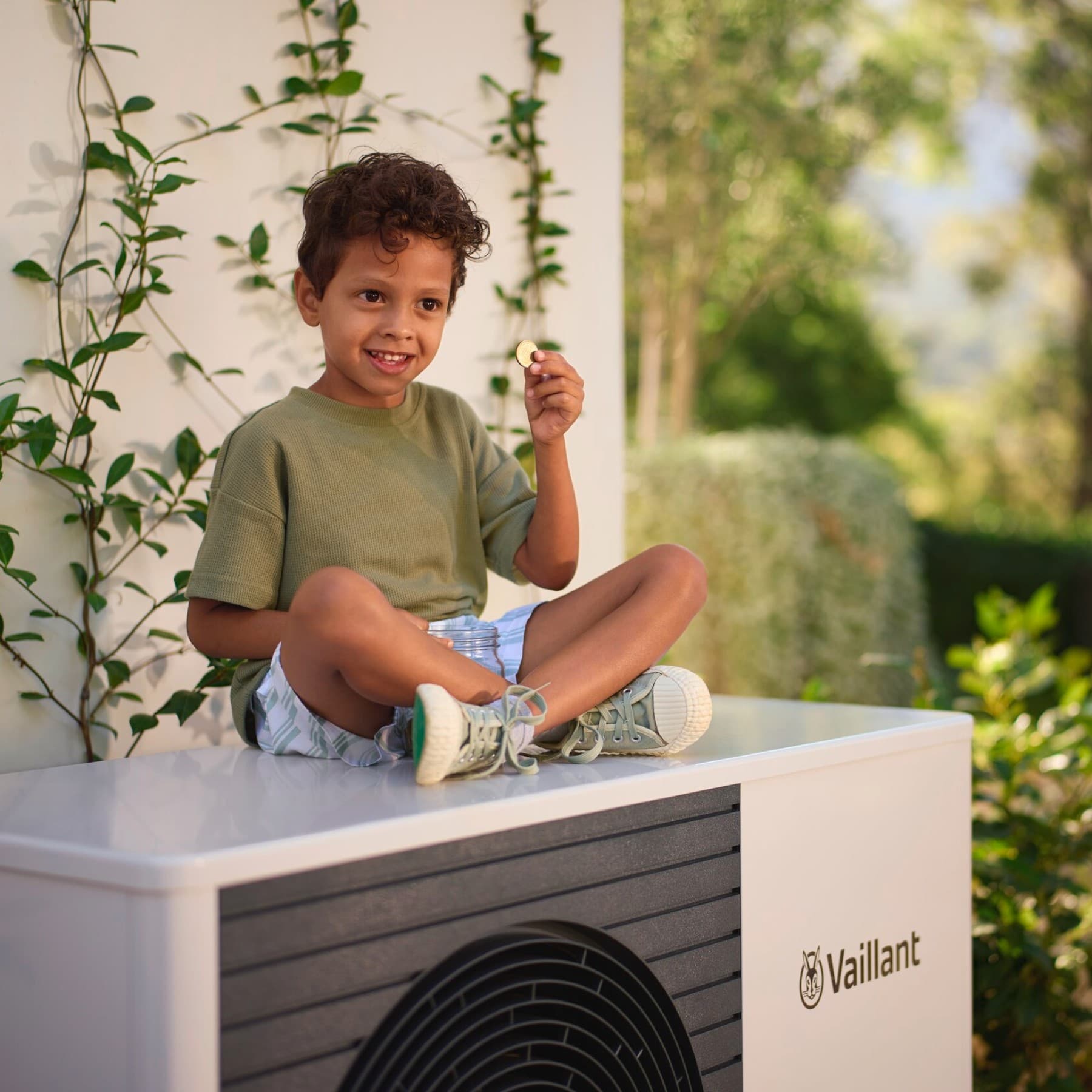 Момче седи на топлинска пумпа во градината држејќи паричка