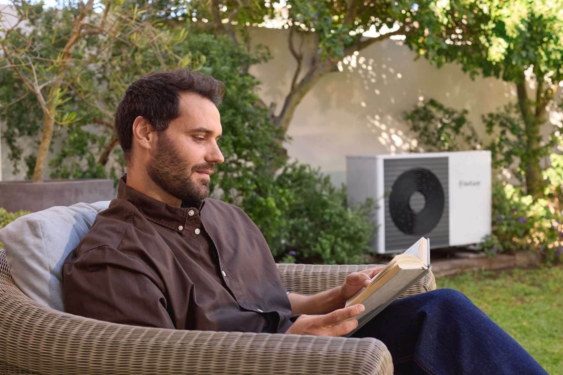 Човек чита книга во градина пред топлинска пумпа