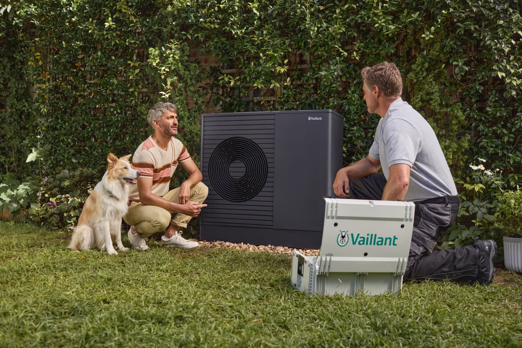 Инсталатер седи клекнат пред топлинска пумпа и му објаснува нешто на еден човек.